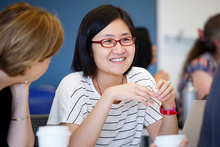 Two women sit at a table with coffee cups talking.